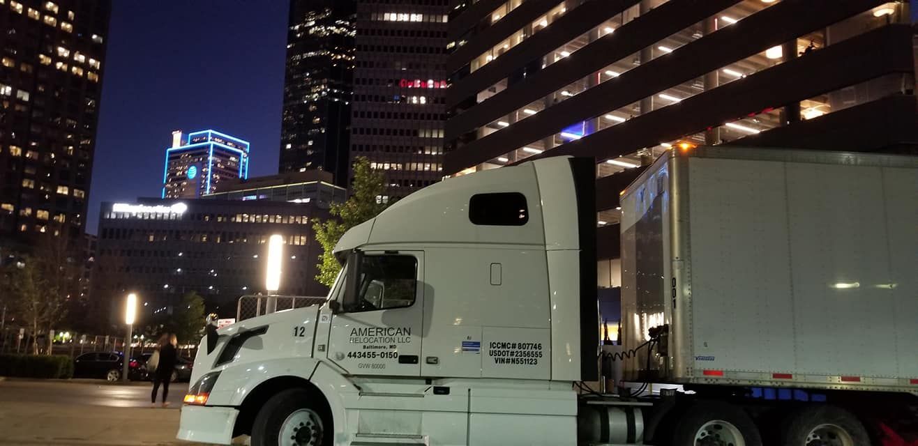 A white semi-truck parked in a city parking lot at night, with illuminated office buildings in the background.