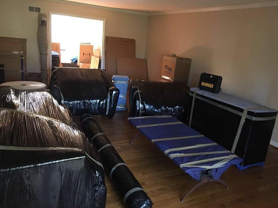 A room filled with furniture wrapped in black plastic and cardboard boxes, likely in preparation for a move. A folded blue cot is also present.