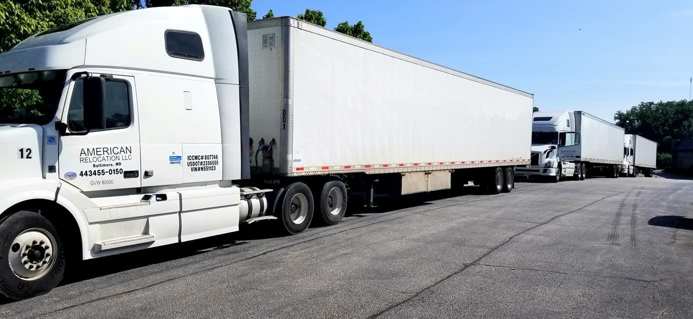 White semi-trucks parked on a paved road under a clear sky. Trees visible in the background.