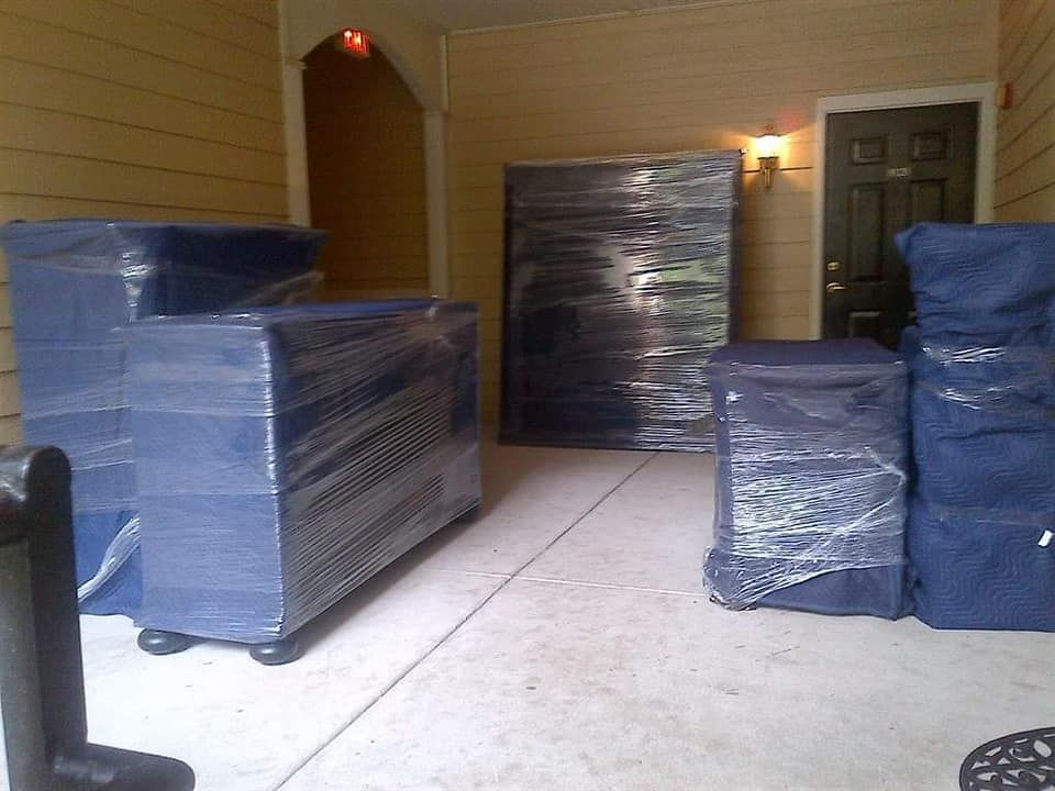 Furniture wrapped in blue pads and plastic wrap on a concrete patio, next to a door and exterior wall.