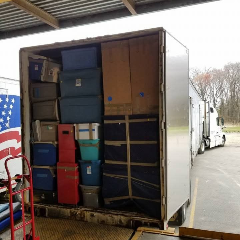 A person in a U.S. military uniform holds a small model house in their hands in front of an American flag.