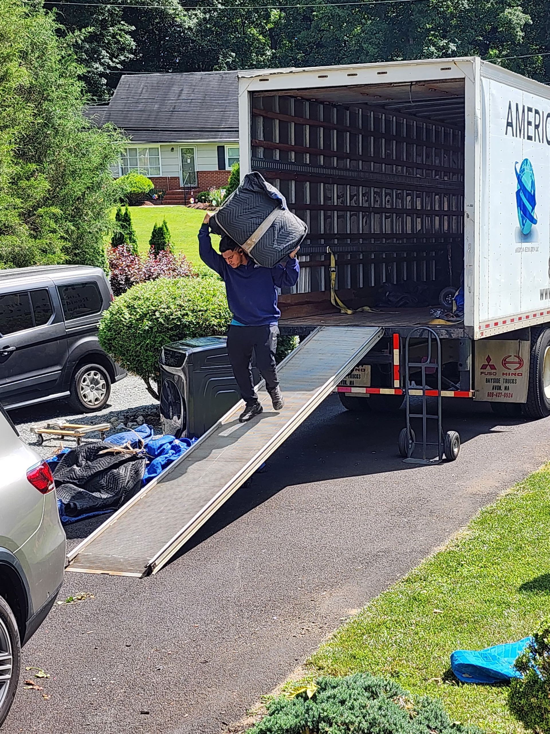 Piano wrapped in plastic and cardboard, secured with straps, being moved. A mover is in the background carrying a piece of furniture.