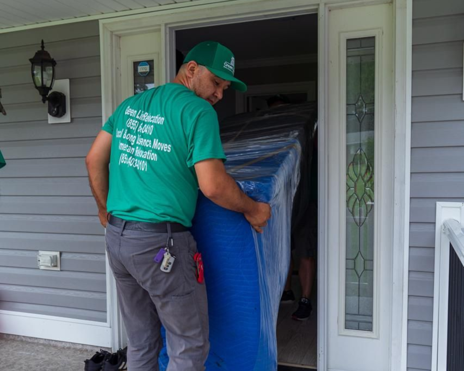 Movers carrying a large, blue wrapped item through a doorway. Person in green shirt, gray pants.