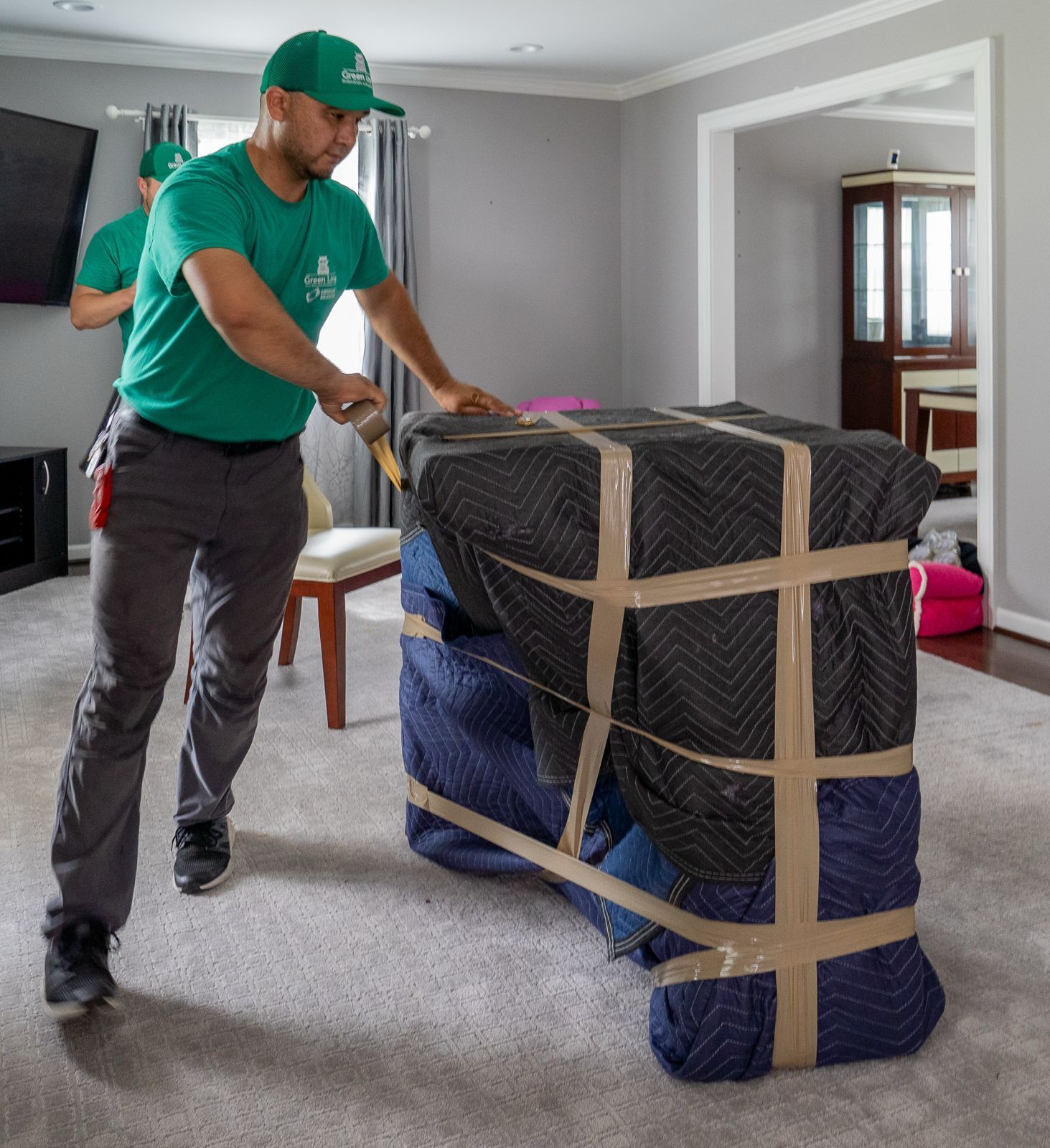 Movers wrap furniture in blankets, securing with tape, inside a home. One pulls the wrapped object.