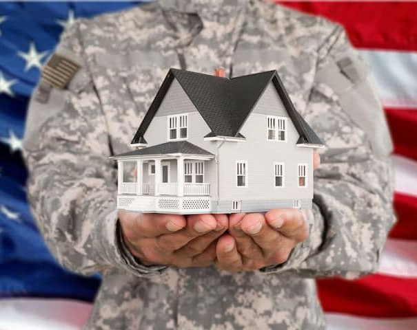 A person in a U.S. military uniform holds a small model house in their hands in front of an American flag.