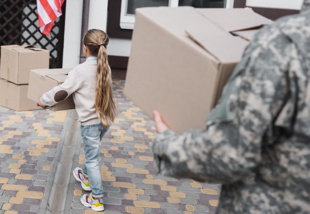 A person in camouflage uniform and a child carry cardboard boxes toward a house with an American flag.