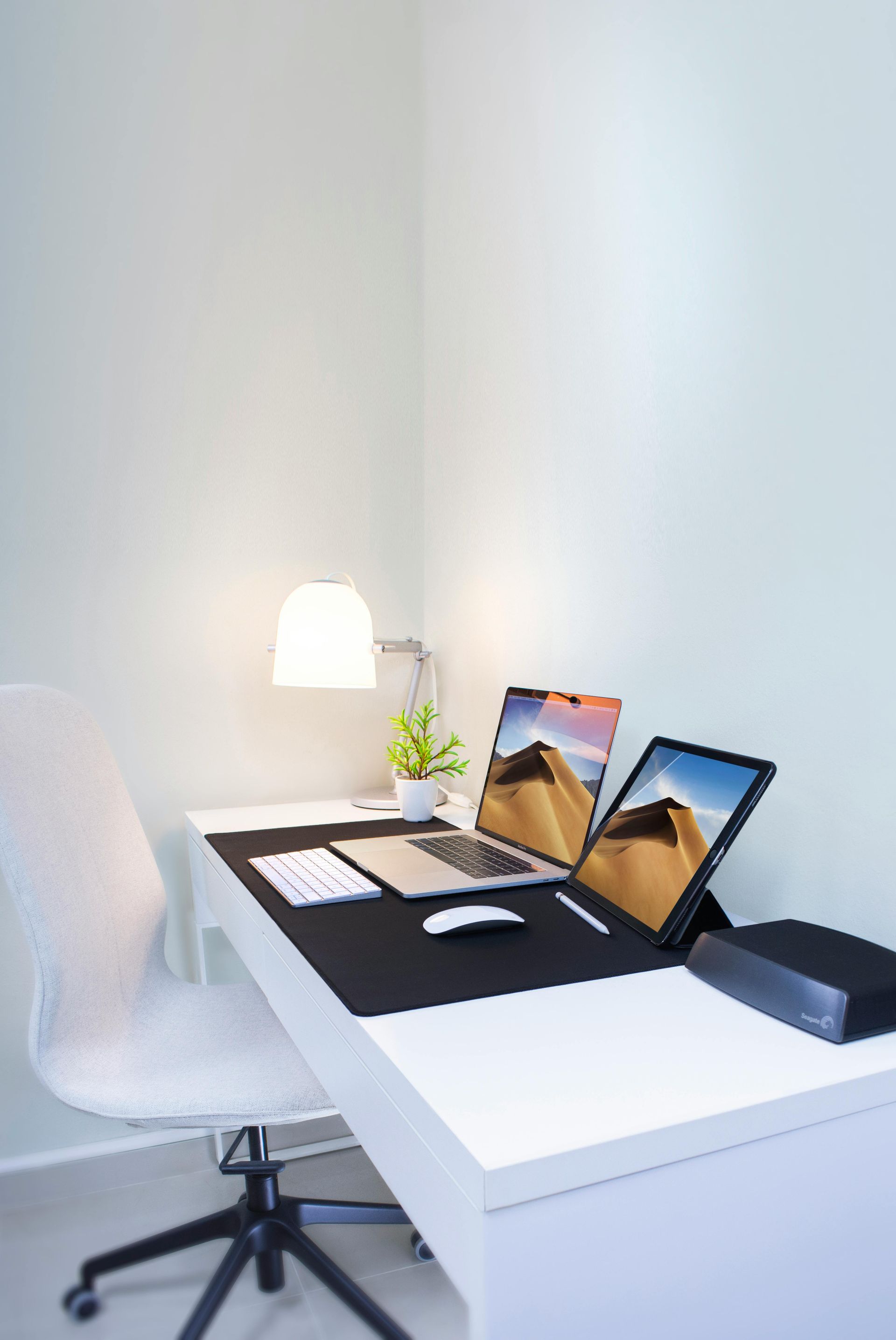 Modern white desk in a corner with laptop, tablet, lamp, and chair; minimalist workspace.