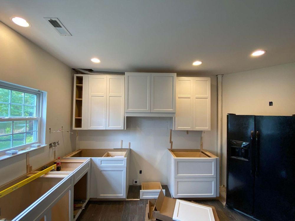 A kitchen under construction with white cabinets and a black refrigerator.