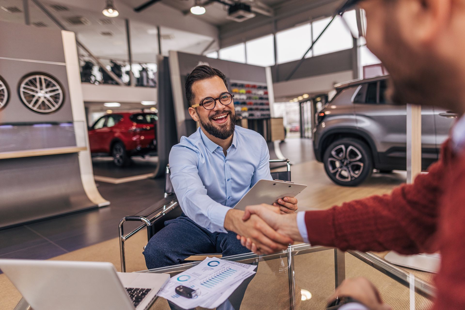Car salesman shaking hands with a customer at a dealership. The salesman is smiling.