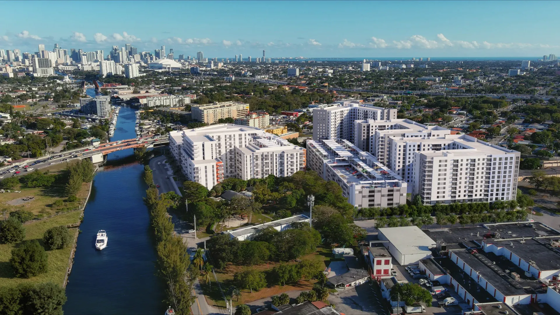 Aerial view of a large white apartment complex beside a canal with a distant city skyline.