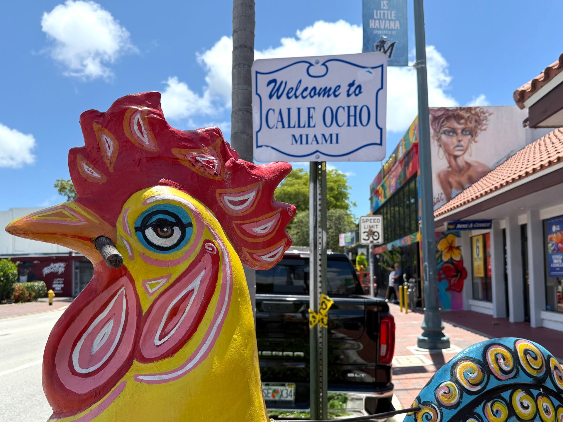 Colorful rooster sculpture welcomes visitors to Calle Ocho, Miami, with street sign and shops visible.