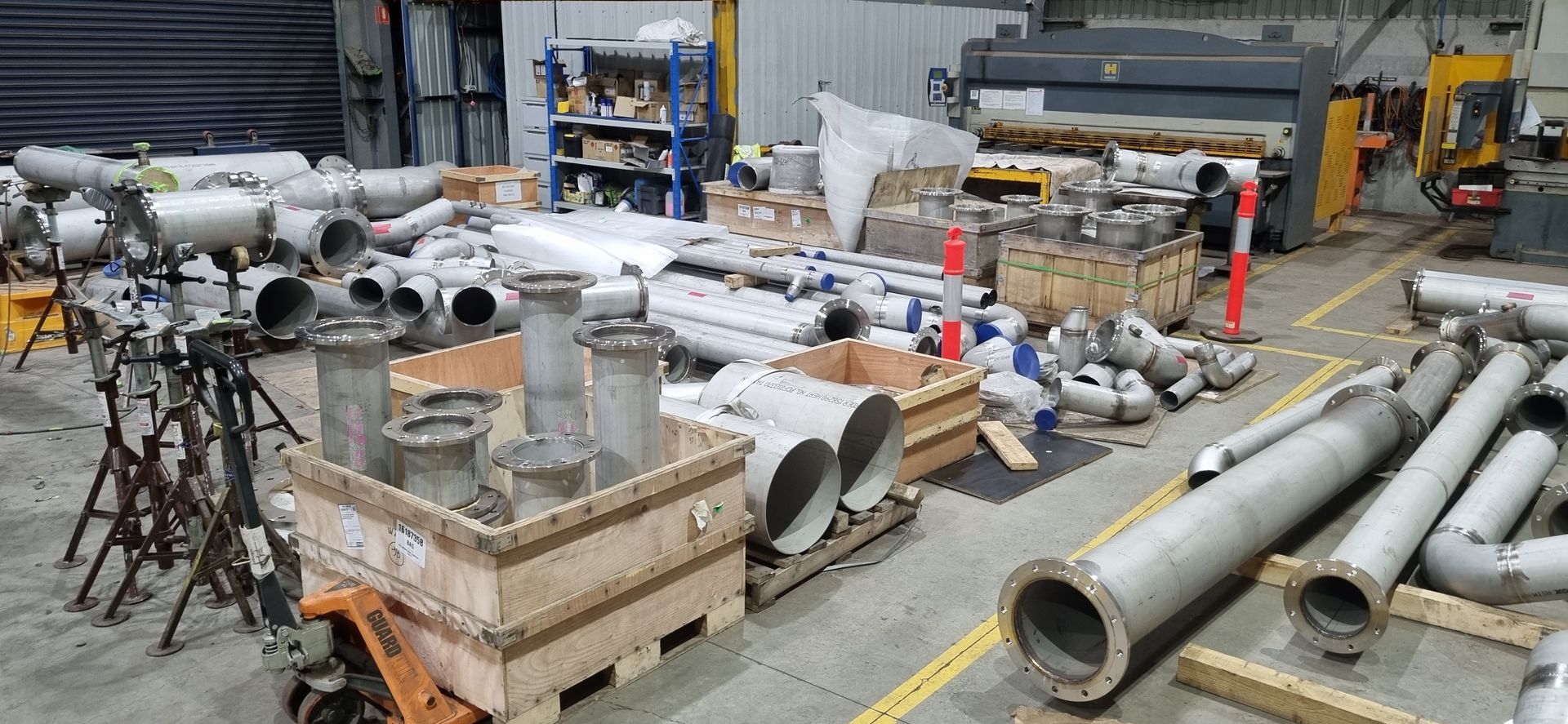 A workshop floor filled with stainless steel pipes, fittings, and industrial materials in crates and on racks.