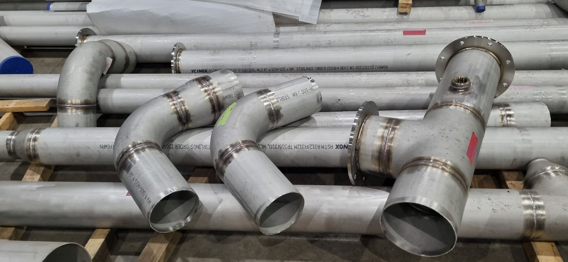 Stainless steel industrial pipes, elbows, and a flanged T-joint resting on wooden pallets in a workshop. Stainless steel industrial pipes, elbows, and a flanged T-joint resting on wooden pallets in a workshop.
