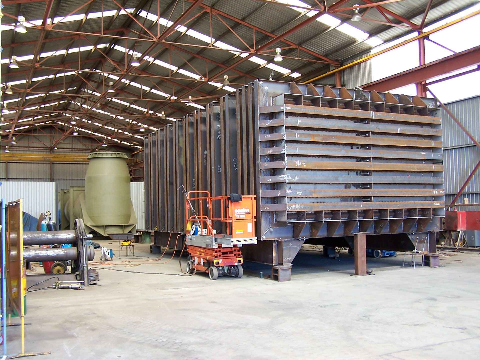 Large metal industrial structure inside a warehouse, likely in construction. Orange lift platform at the front.