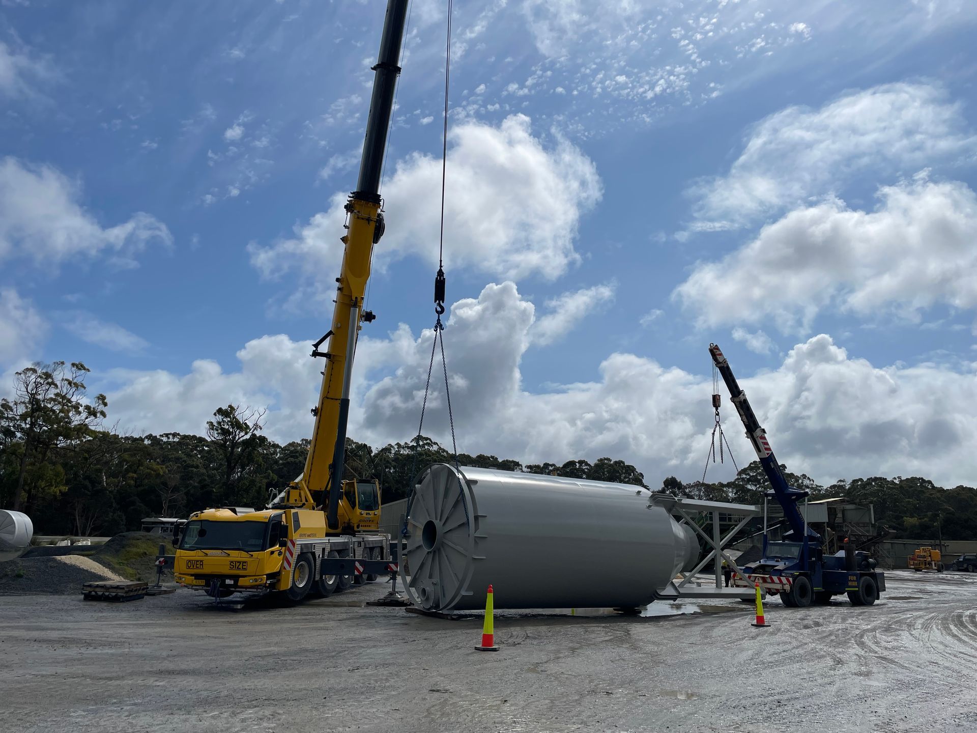 Two cranes lifting a large gray cylindrical structure on a construction site under a cloudy sky.