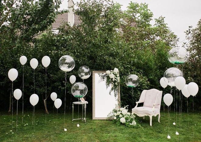 White balloons and clear orbs decorate a garden with a mirror and chair; green foliage in background.