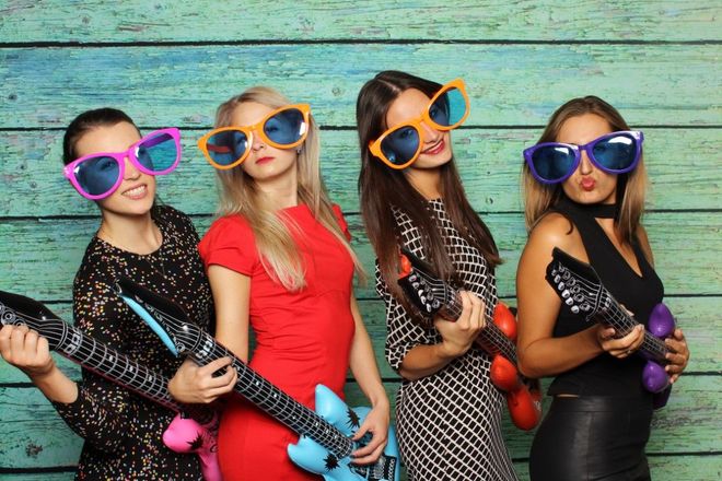 Four women with oversized sunglasses, holding toy guitars, posing in front of a teal wooden backdrop.