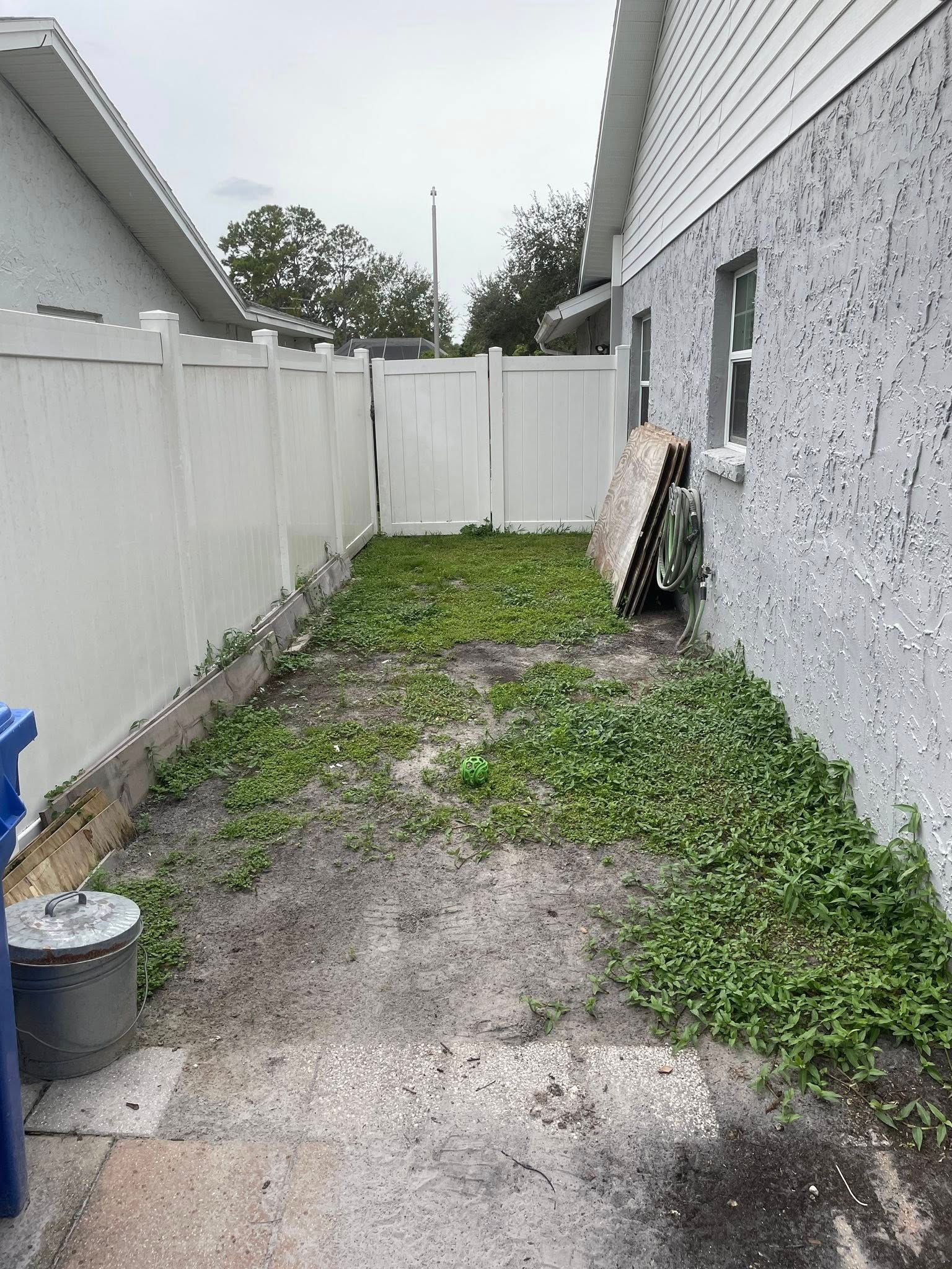 A narrow backyard with a white fence, gray ground, and patches of green weeds next to a light gray building.