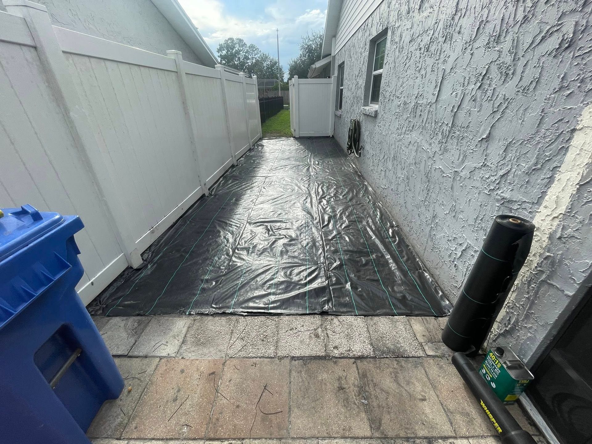 Narrow outdoor area with black tarp on paving stones between a white fence and stucco wall.