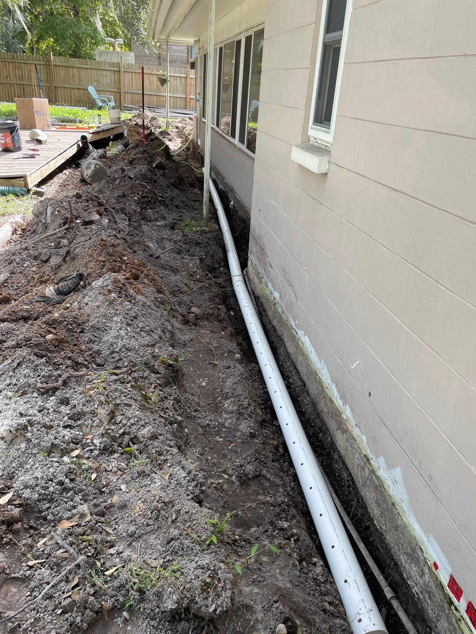 Trench alongside a house with a white perforated pipe. The ground is dark soil.