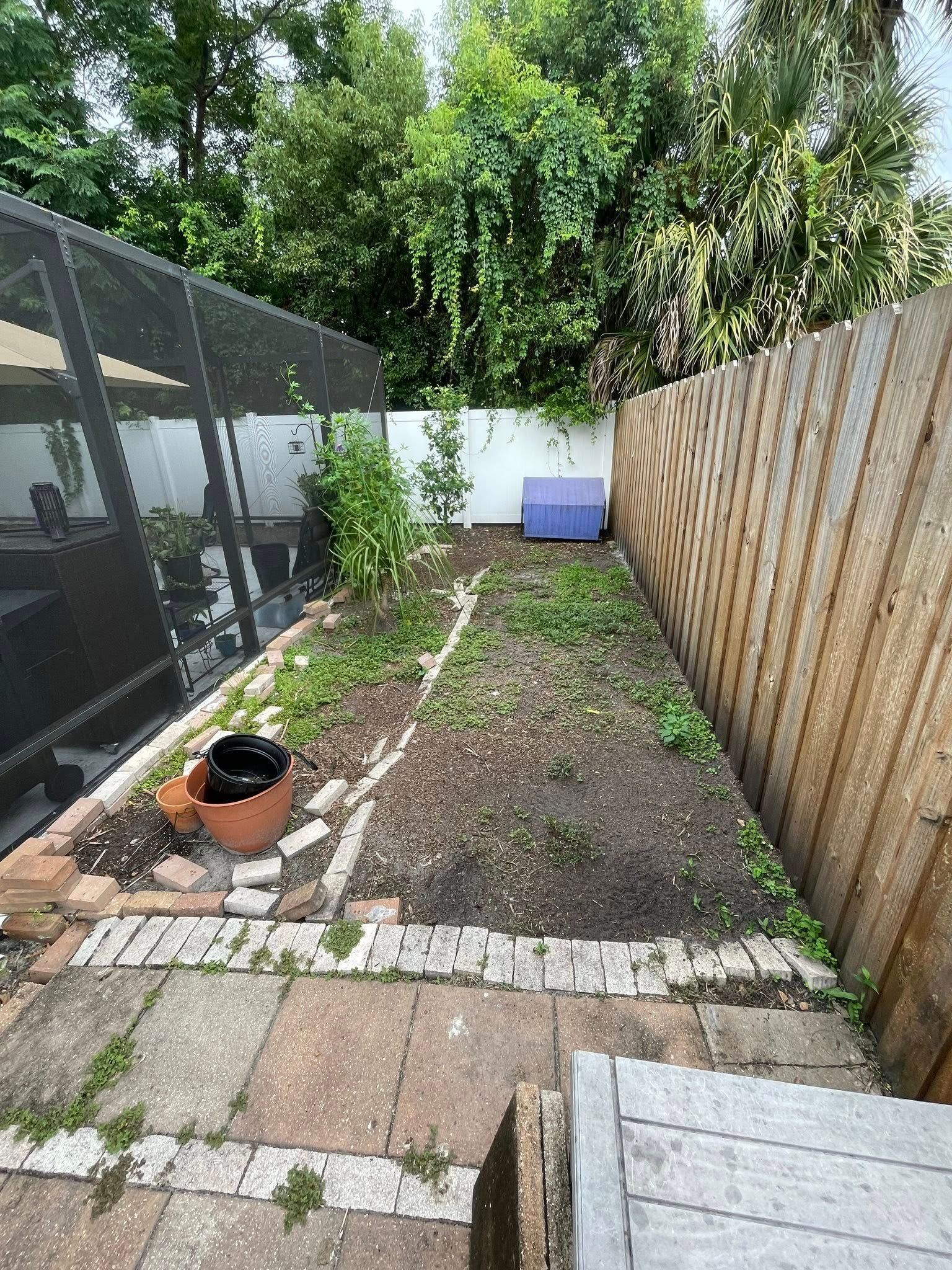 Backyard garden with brick and stone path, fence, and plants. Brown soil and green foliage.