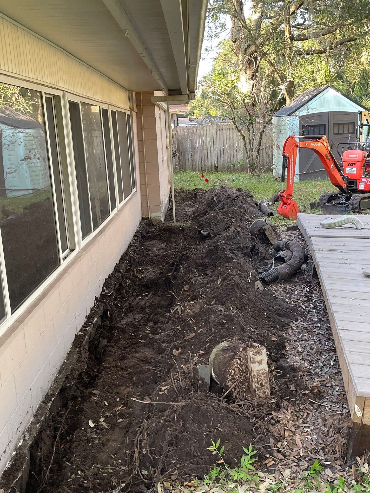 Trench dug alongside a building; mini excavator in background; dirt and debris present.