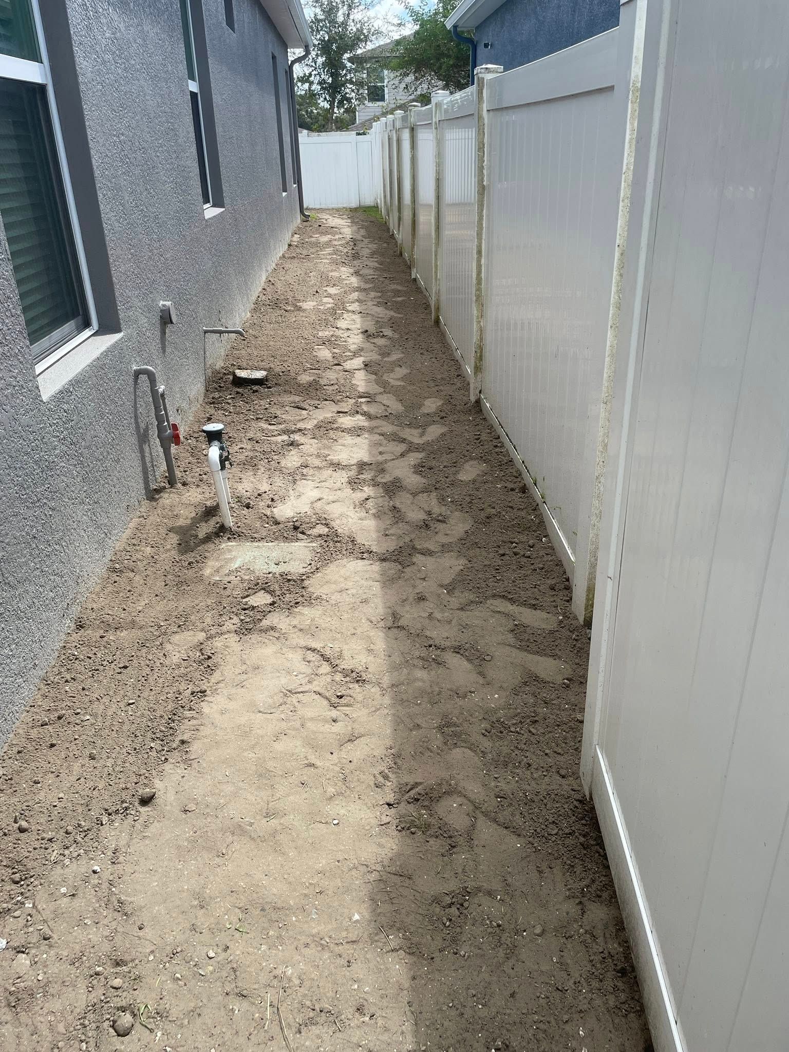 Narrow dirt path between a stucco building and a white fence, in a sunny outdoor setting.