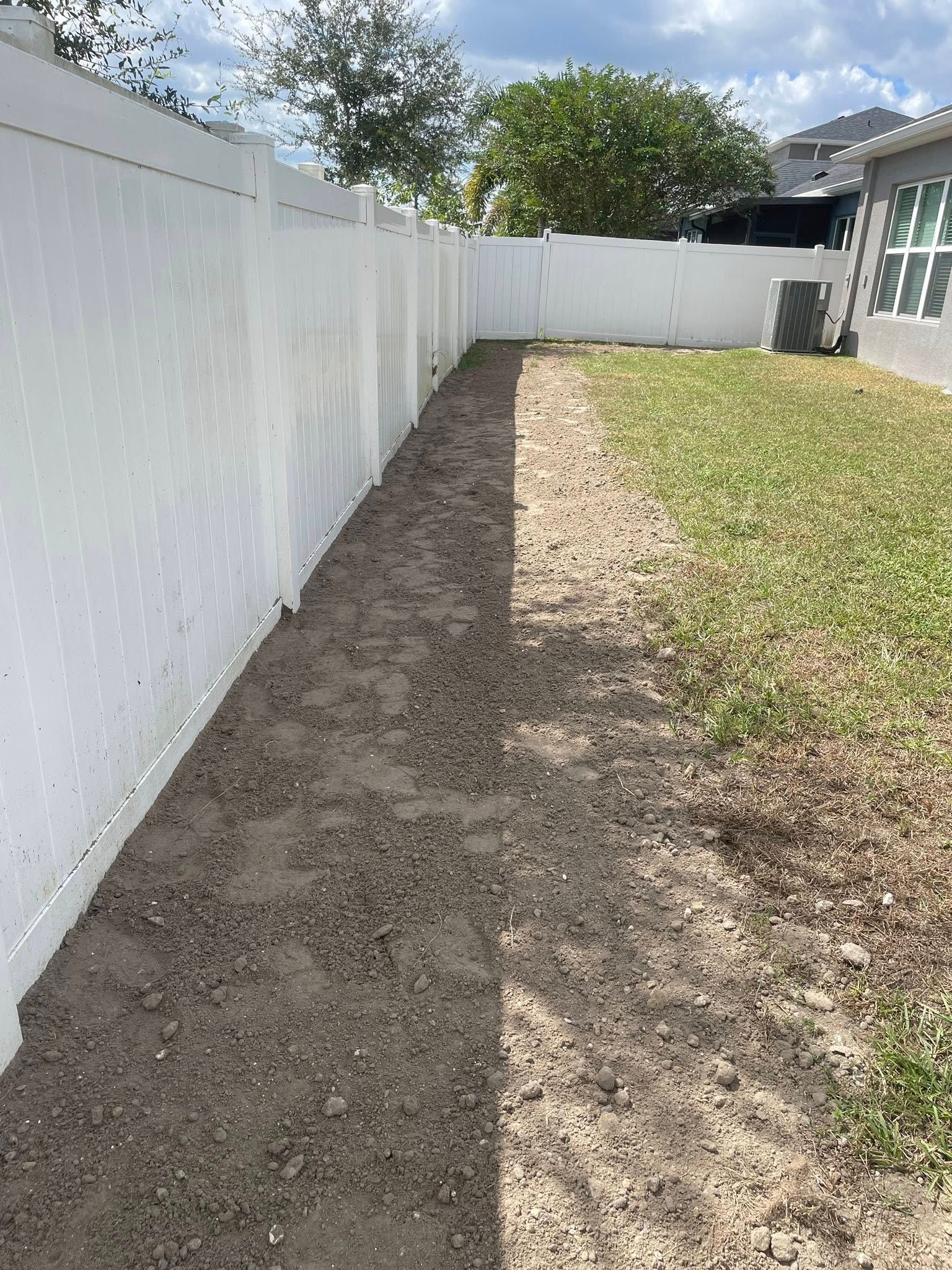 A narrow dirt path next to a white fence in a backyard with green grass.