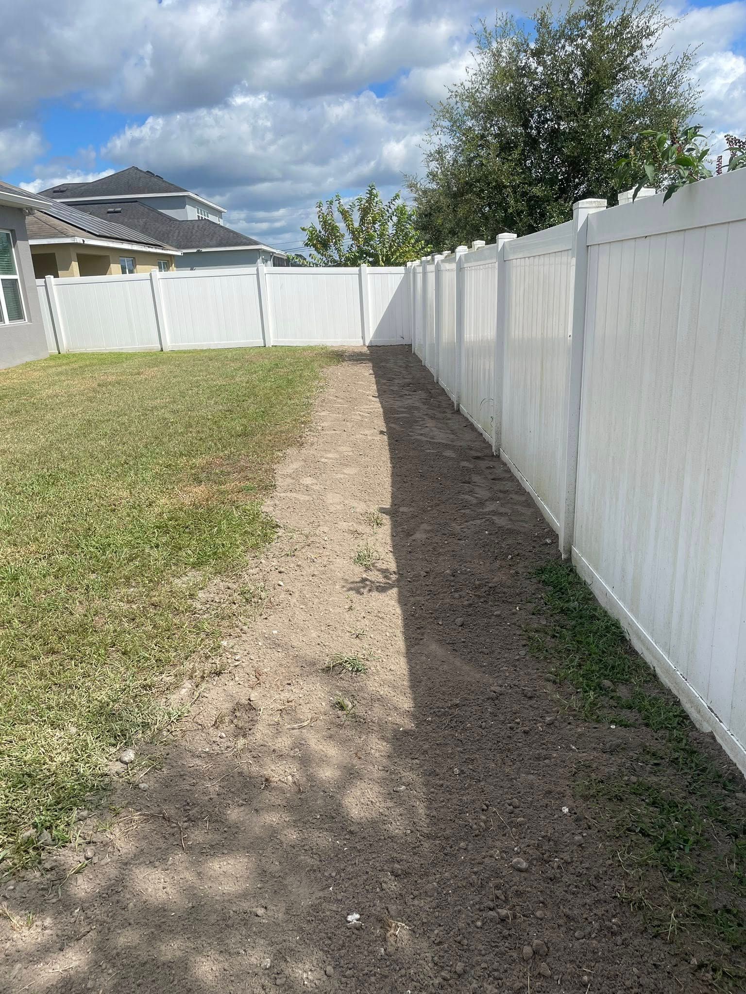 A narrow dirt patch alongside a white vinyl fence in a grassy yard.