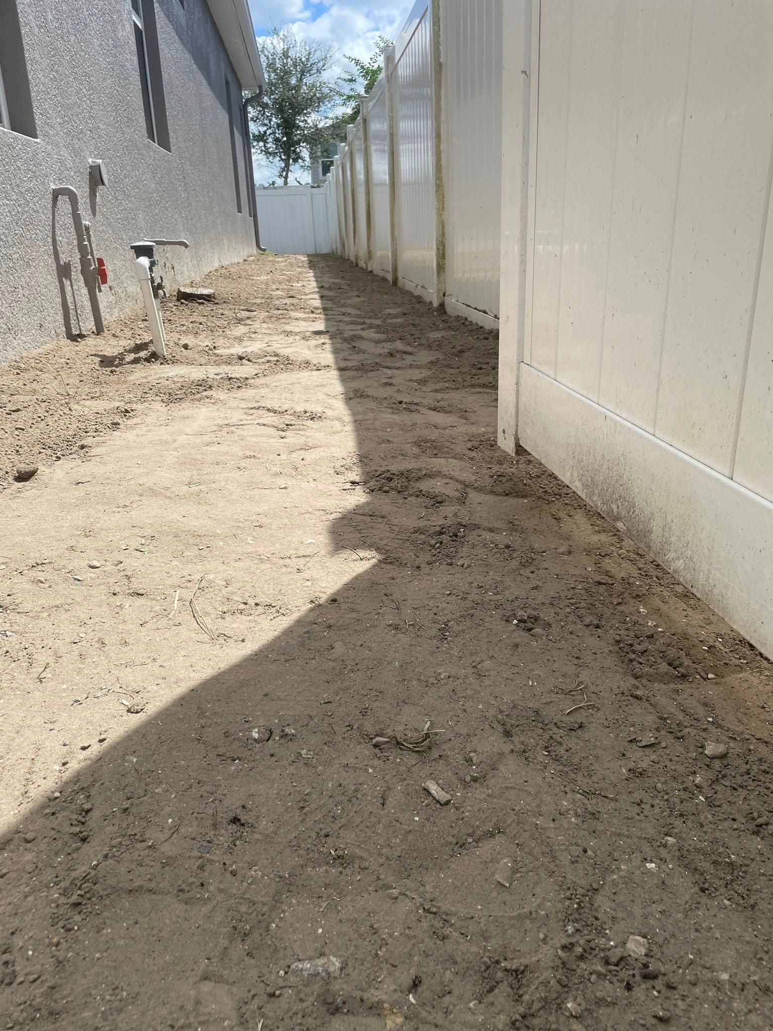 Narrow dirt pathway between a white fence and building; bright sunlight.
