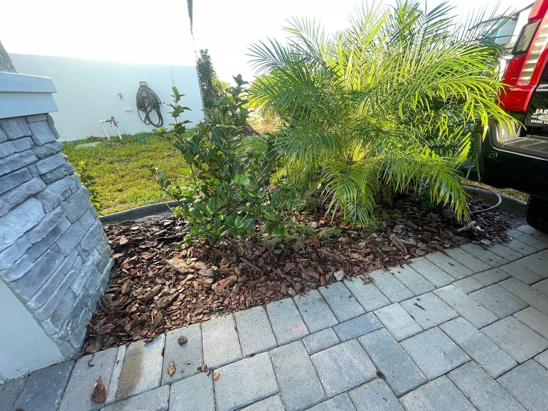 Stone column and paved driveway border a landscaped garden bed with mulch and green plants.