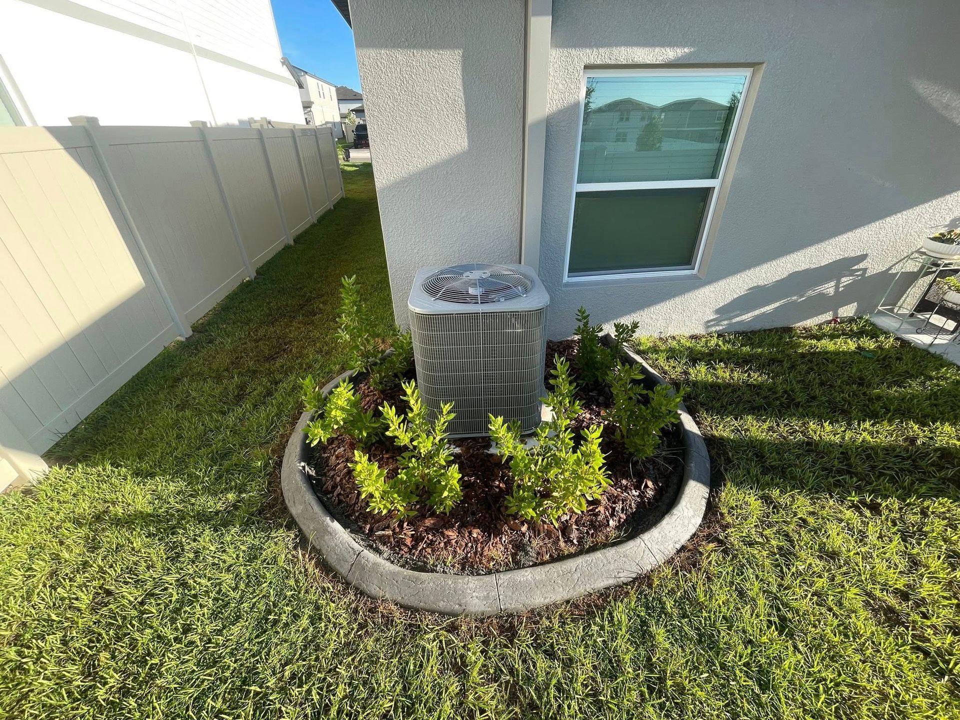 Air conditioner surrounded by plants in a concrete-edged garden bed against a light-colored building.