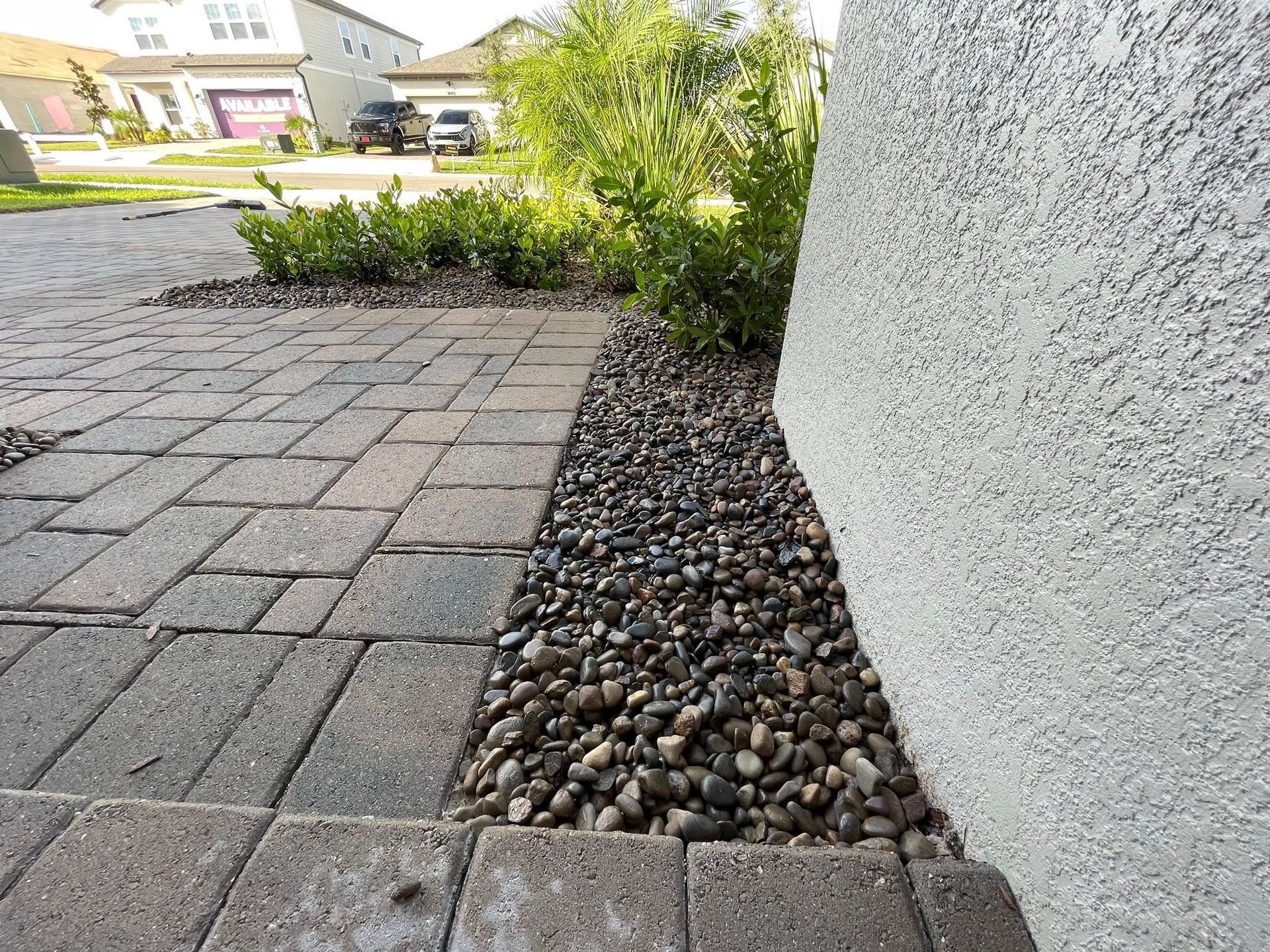 Brick walkway next to a bed of small pebbles and a stucco wall, with green shrubs.