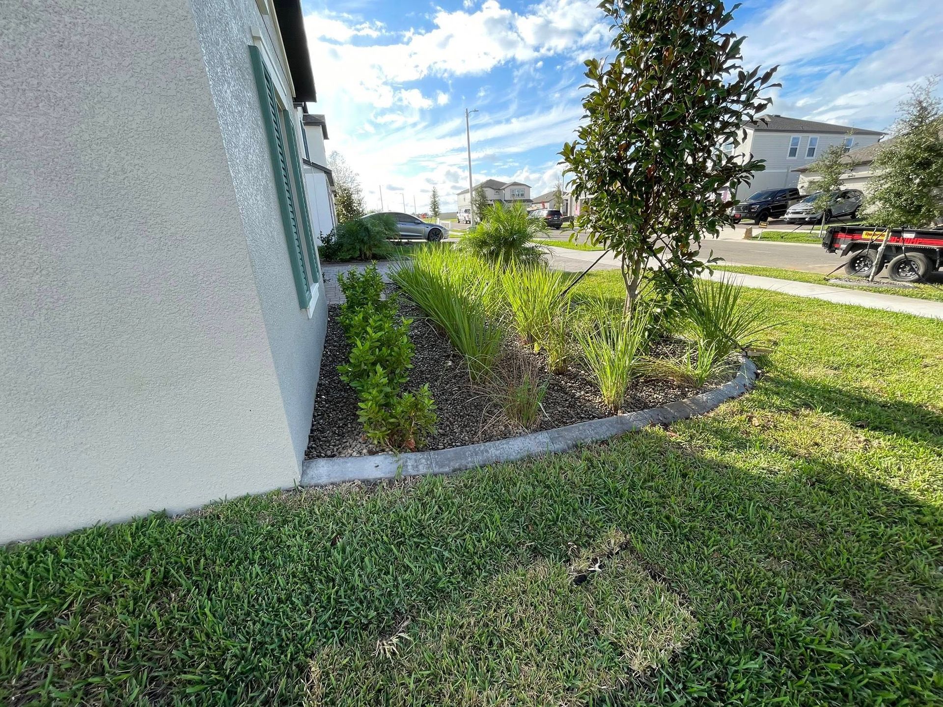 A landscaped yard with a gray concrete border, green plants, and a light-colored house.