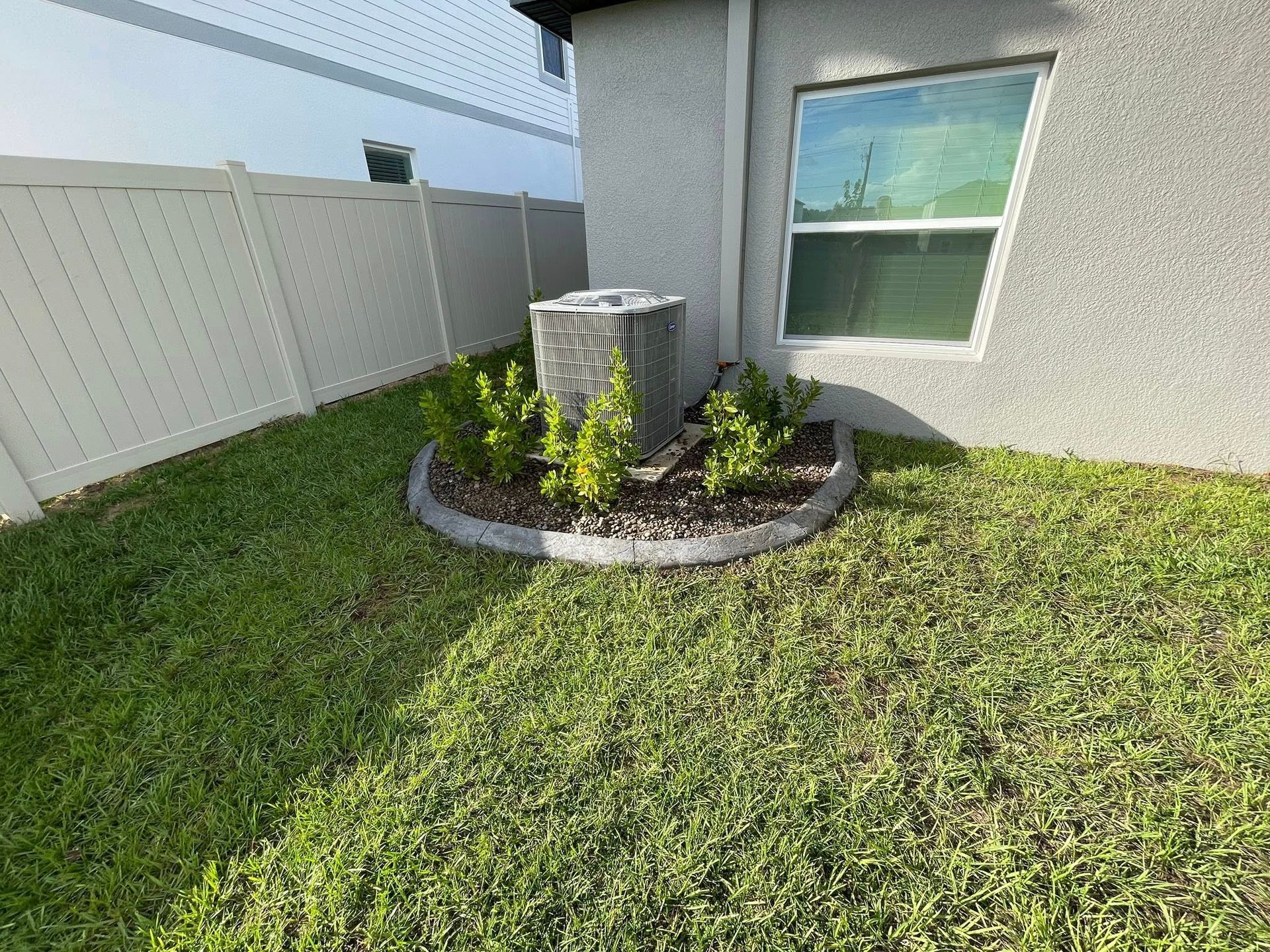 Backyard with a fountain and small garden bed next to a white fence and a house. Green grass covers the ground.