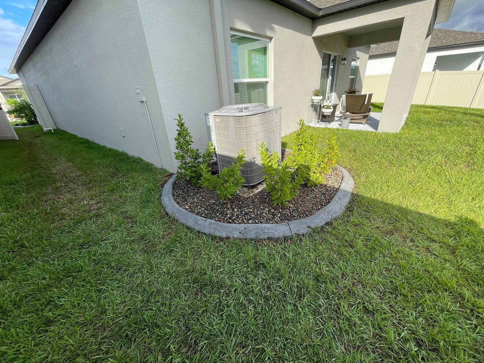 AC unit surrounded by plants in a landscaped bed with a gray concrete border, near a house with patio.