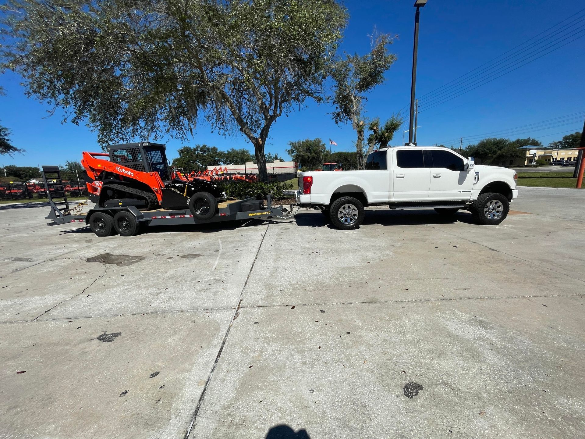 White pickup truck towing a trailer with an orange tractor on a cracked asphalt surface on a sunny day.