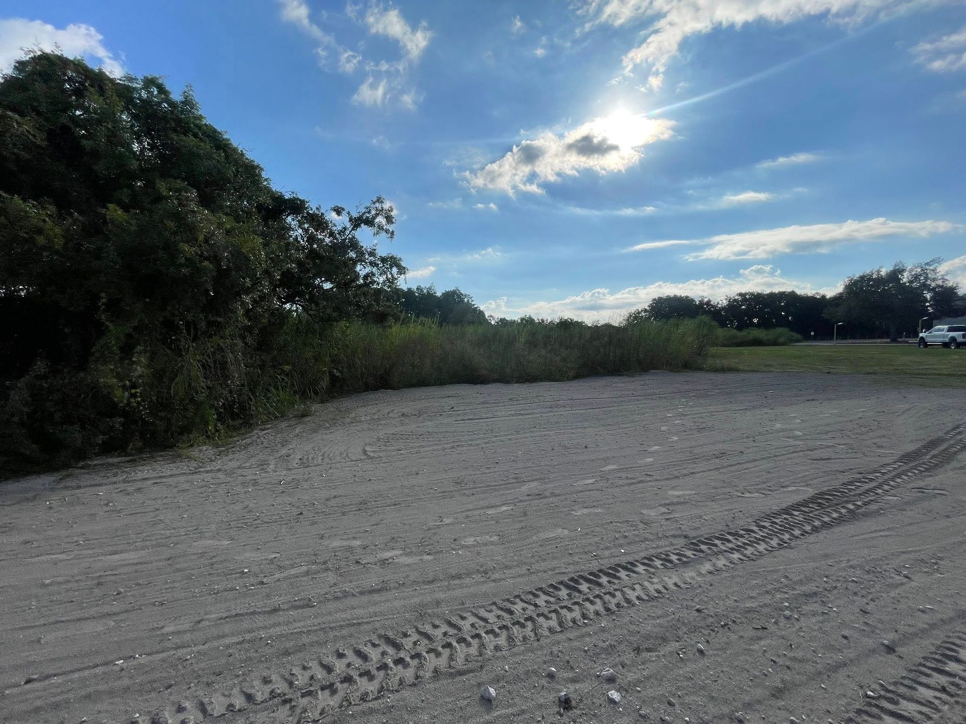 A gravel clearing with tire tracks, bordered by vegetation under a partly cloudy sky.