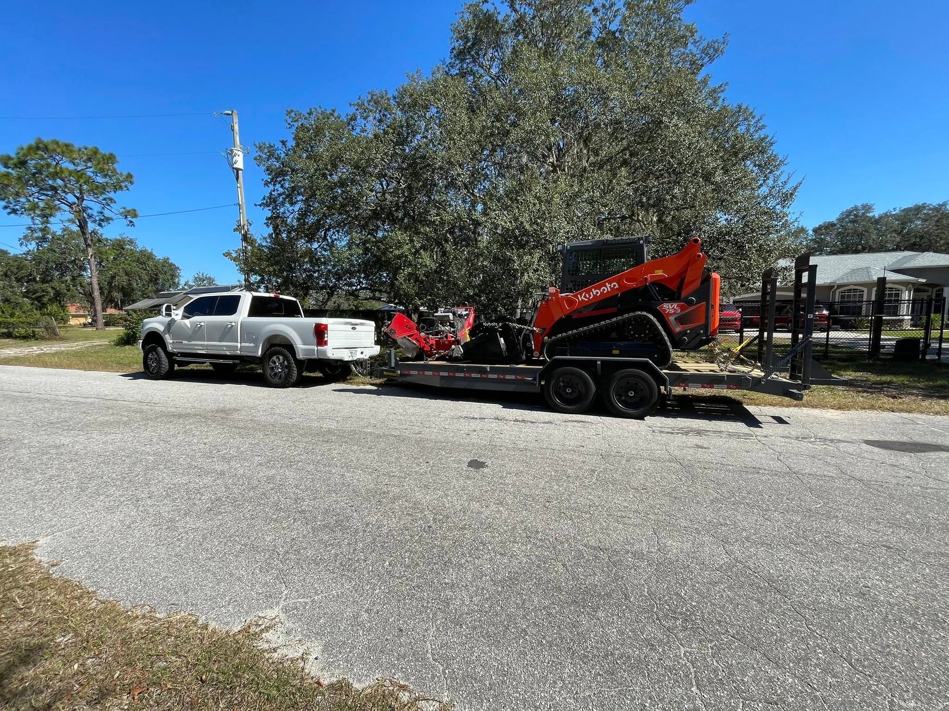 White truck towing a trailer with orange Kubota skid steer on a gravel driveway.