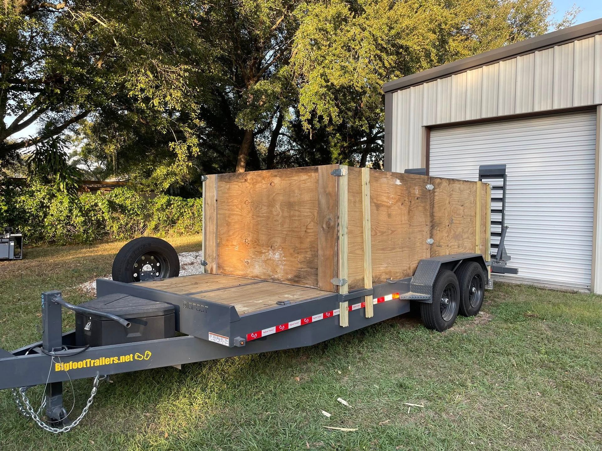 A flatbed trailer with wooden sides parked on grass next to a white garage.