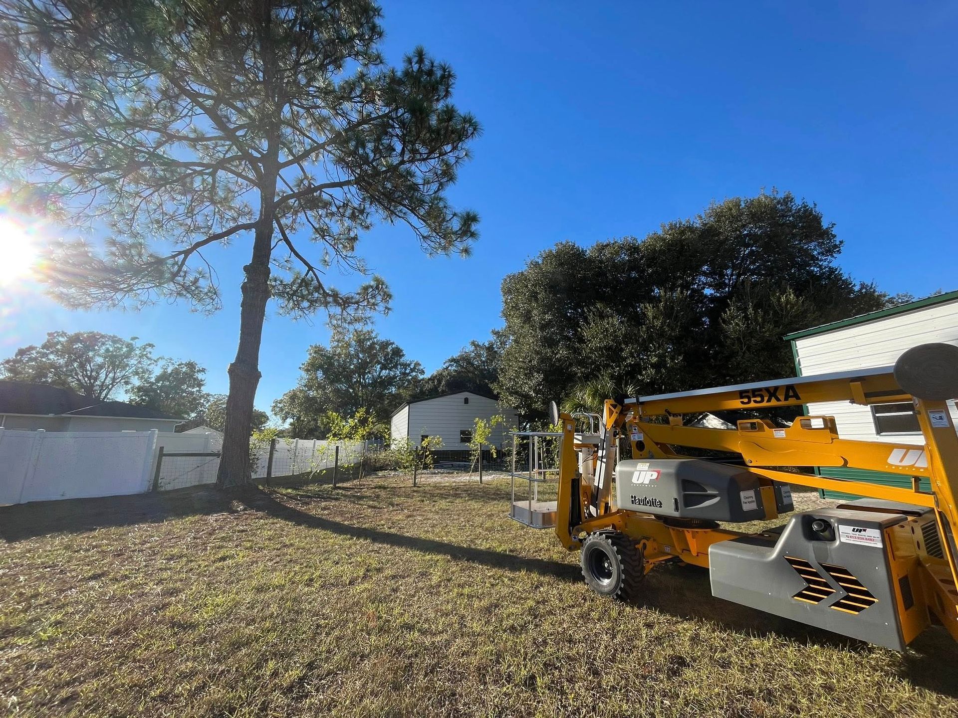 Yellow lift equipment in a yard, tree and house visible under a bright blue sky.