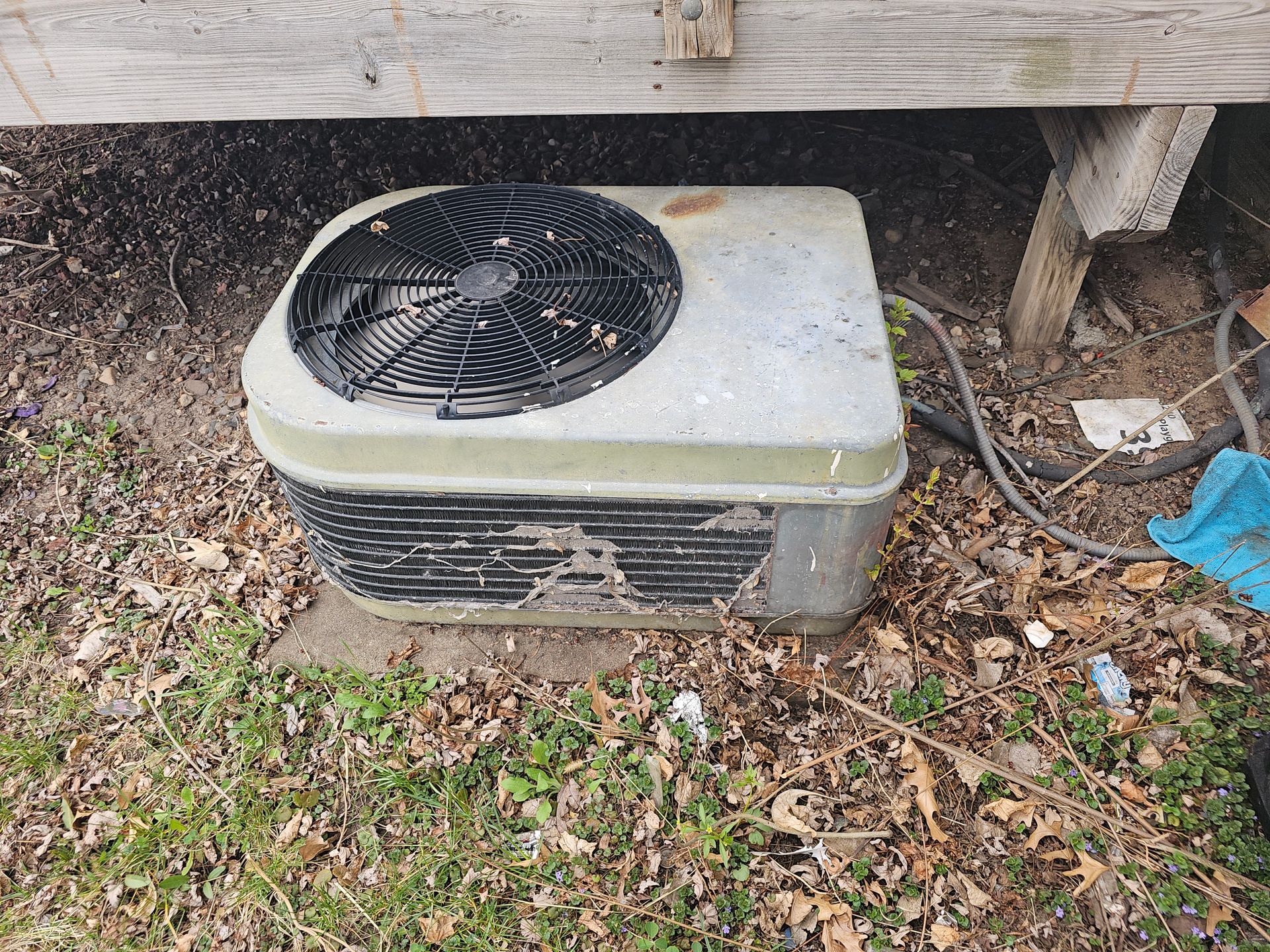 An air conditioner is sitting on the ground under a wooden fence.