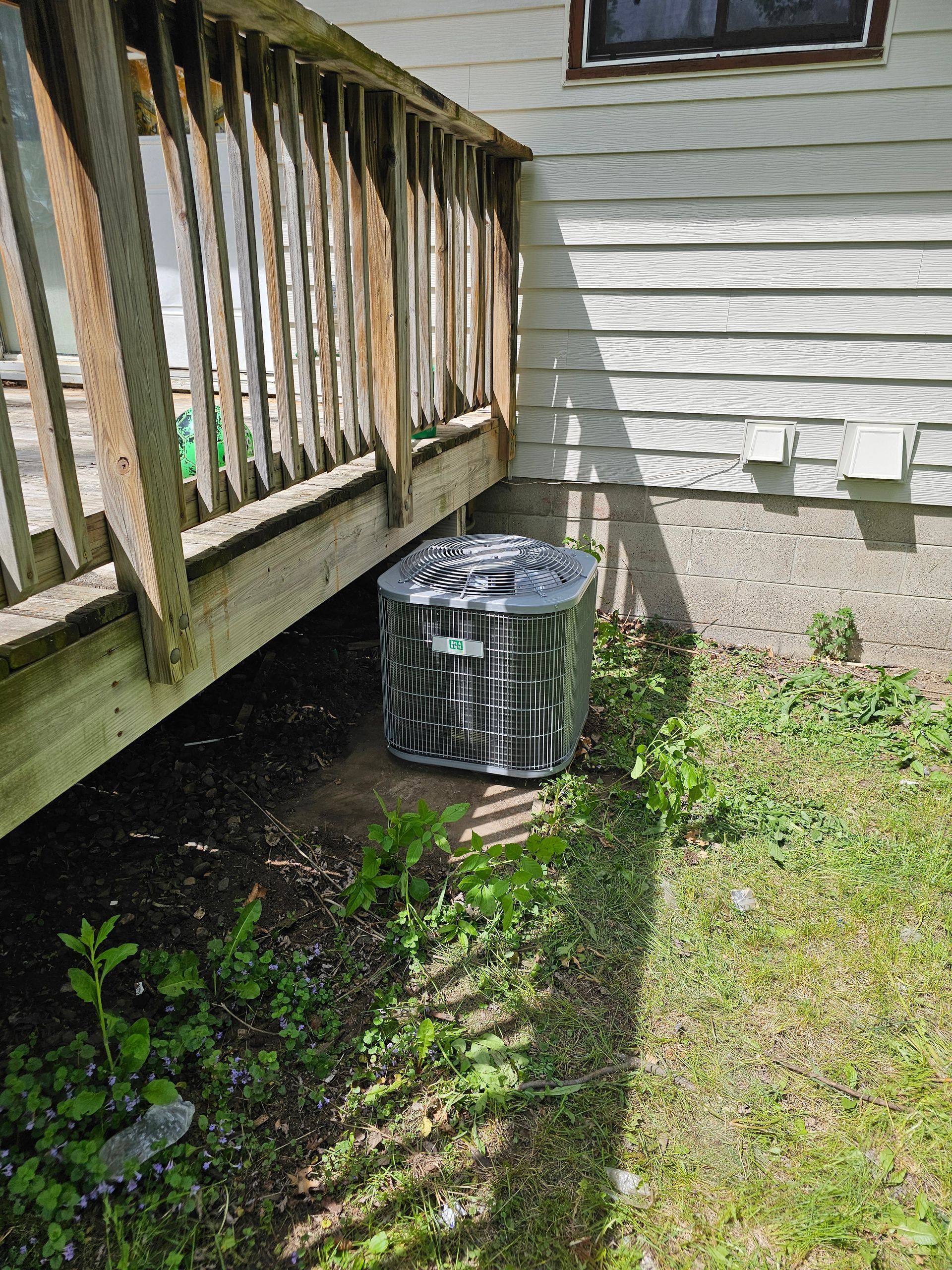An air conditioner is sitting under a deck next to a house.