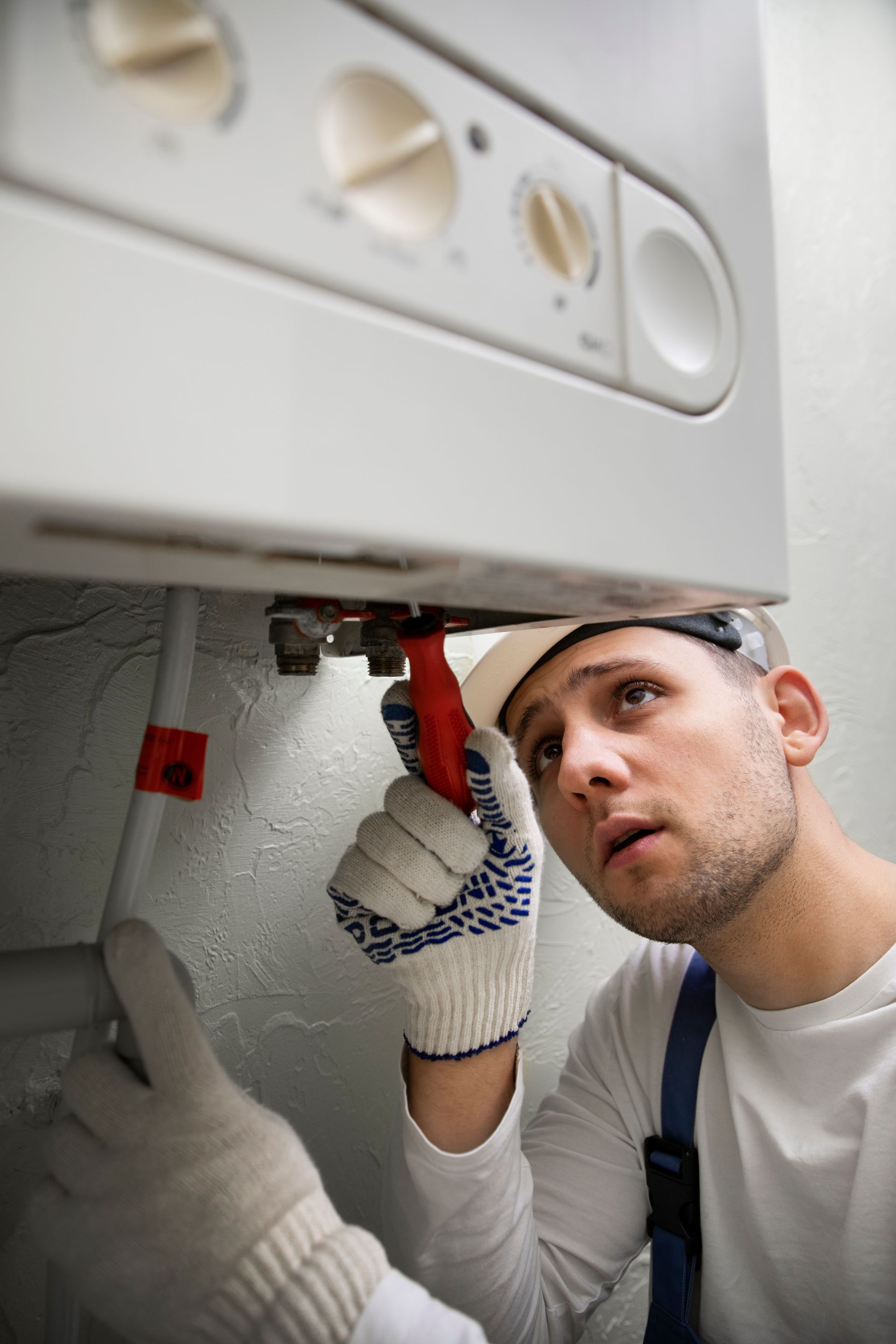 Plumber using a wrench on a boiler, wearing gloves and a hard hat, looking up with a focused expression.