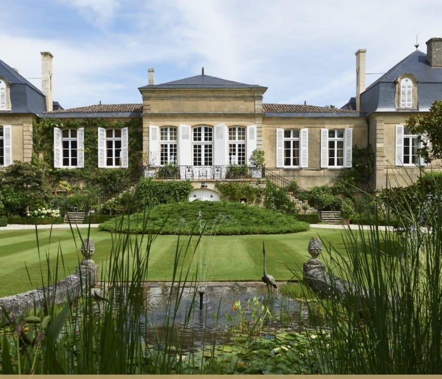 Une berline Mercedes noire est garée devant une grande maison beige entourée d'arbres.