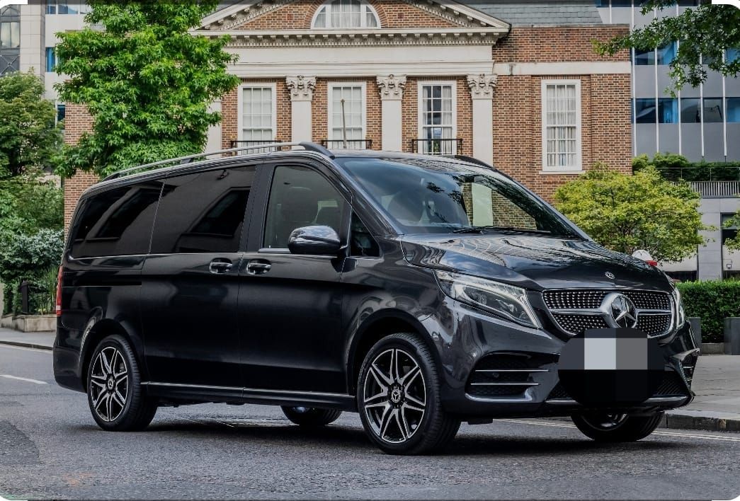 Une camionnette Mercedes-Benz noire est garée dans une rue, devant un bâtiment en briques comportant plusieurs fenêtres.