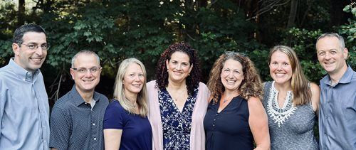 A group of seven people smiling in front of a green leafy background.