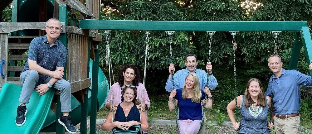 Group of people on a playground; some on swings and others standing near a slide. Outdoors, smiles.