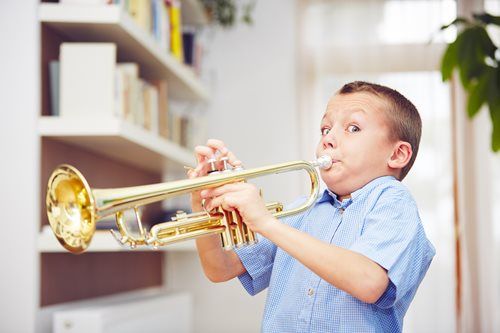 Boy playing a trumpet indoors, eyes wide, cheeks puffed out, near a bookshelf.