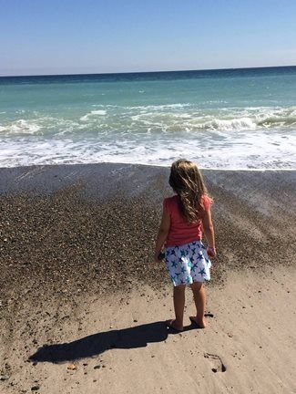 Girl standing on beach, looking at ocean. Blue-green water, sunny day.